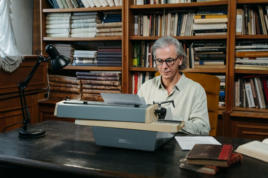 Elderly writer using a typewriter in a vintage library filled with bookshelves.